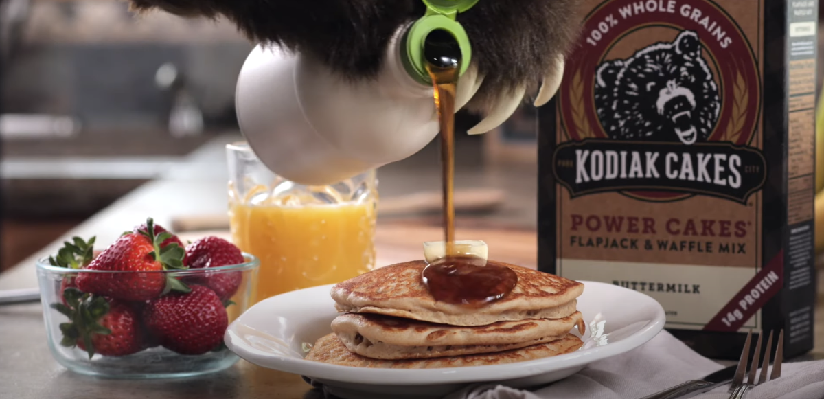 Box of Kodiak cake mix, strawberries, juice, and pancakes being drizzled with syrup.