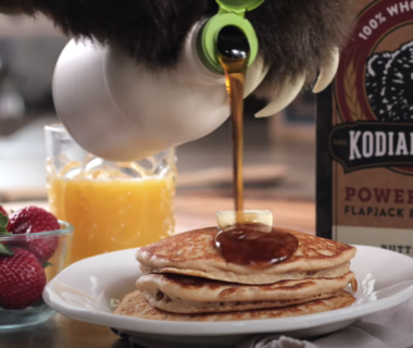 Box of Kodiak cake mix, strawberries, juice, and pancakes being drizzled with syrup.