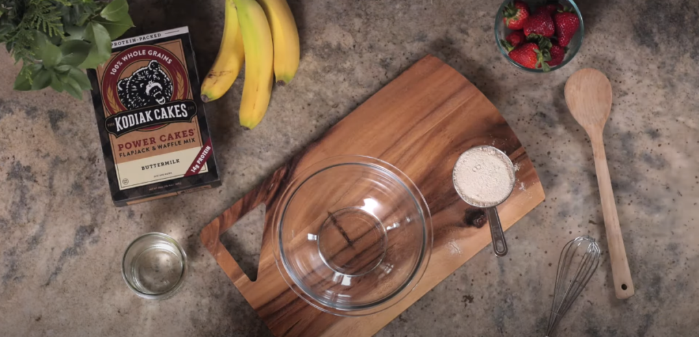 Kodiak cake box, clear bowl on wooden chopping board, measuring spoon with powder, whisk, and strawberries on kitchen countertop.