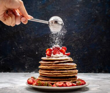 a front view of yummy and round pancakes with cream, powdered sugar, and red strawberries