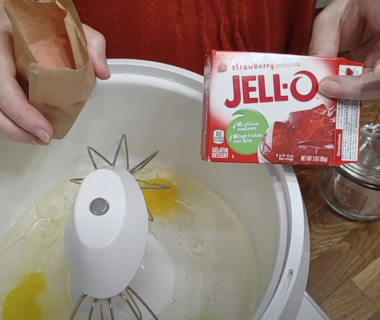 Hand holding a box of Jello while a stand mixer contains eggs.