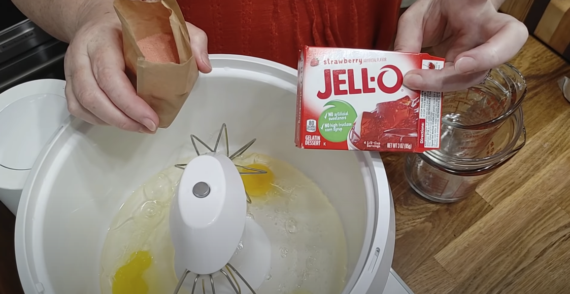 Hand holding a box of Jello while a stand mixer contains eggs.