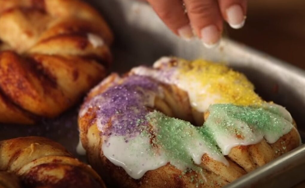 a close depiction of a mini-sized king cake and a hand adding sprinkles on the cake